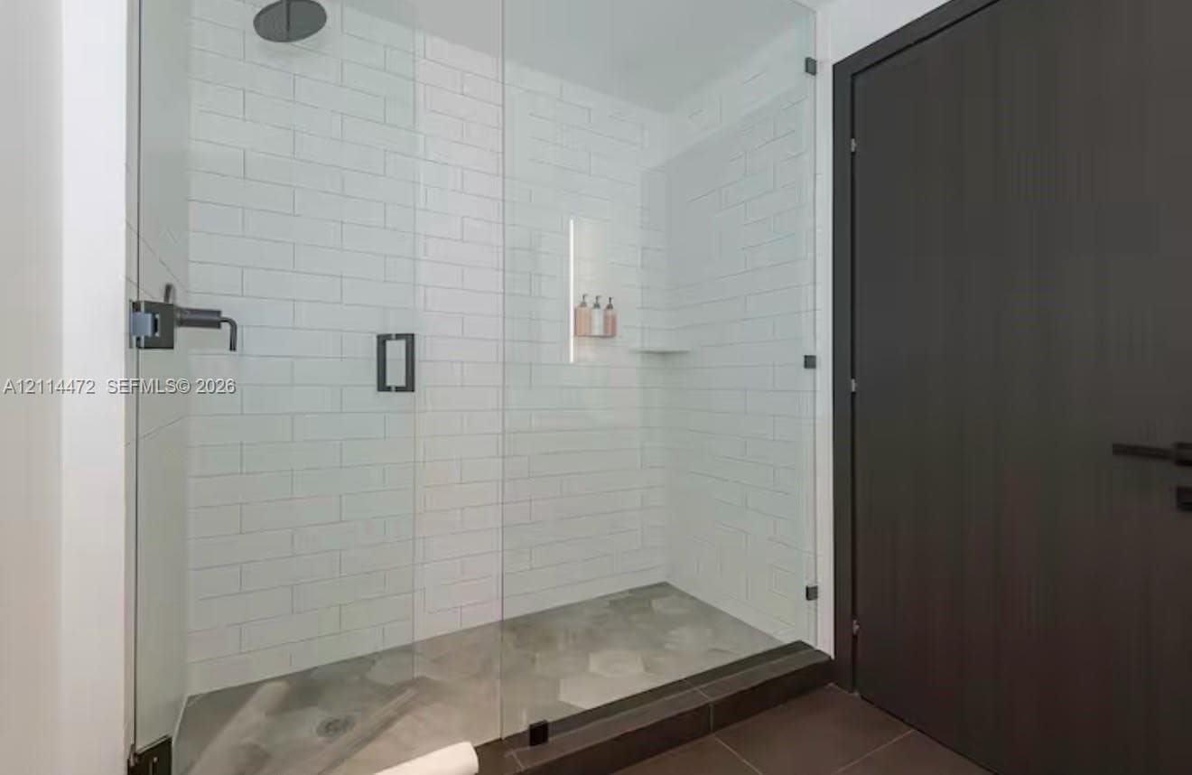 Walk-in shower featuring a frameless glass enclosure, white subway tile surround, hexagonal tile flooring, matte black rainfall showerhead, and integrated recessed shelving