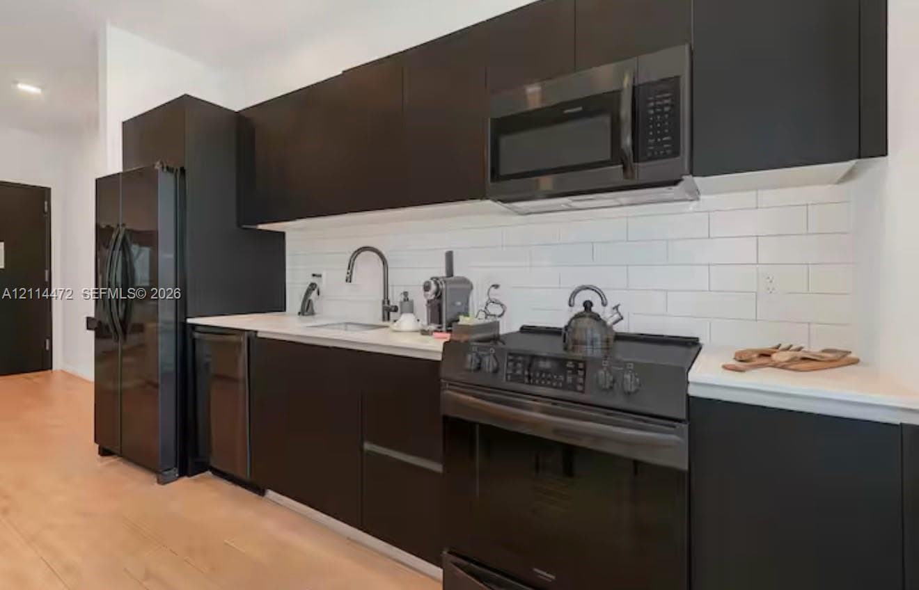 Modern kitchen featuring matte black flat-panel cabinetry, white solid surface countertops, white subway tile backsplash, and a suite of black appliances