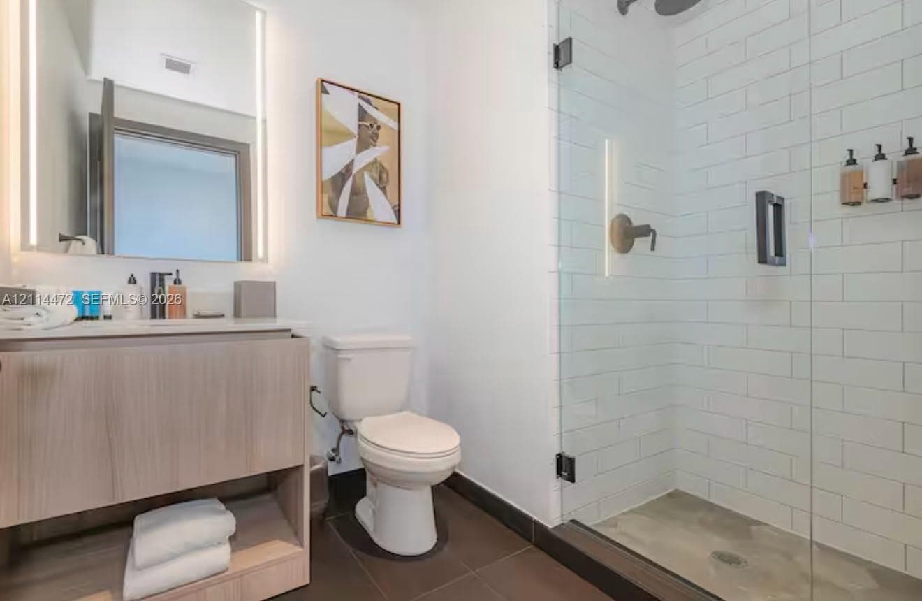 Wood-finish floating vanity with an integrated countertop, illuminated wall-mounted mirror, walk-in shower with white subway tile surround, and large-format tile flooring