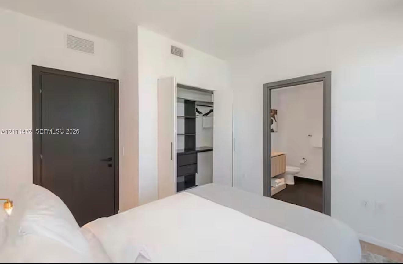 Neutral-toned bedroom featuring a dark solid-core door, built-in closet with integrated shelving, and a doorway showcasing a vanity with a light-finish cabinet