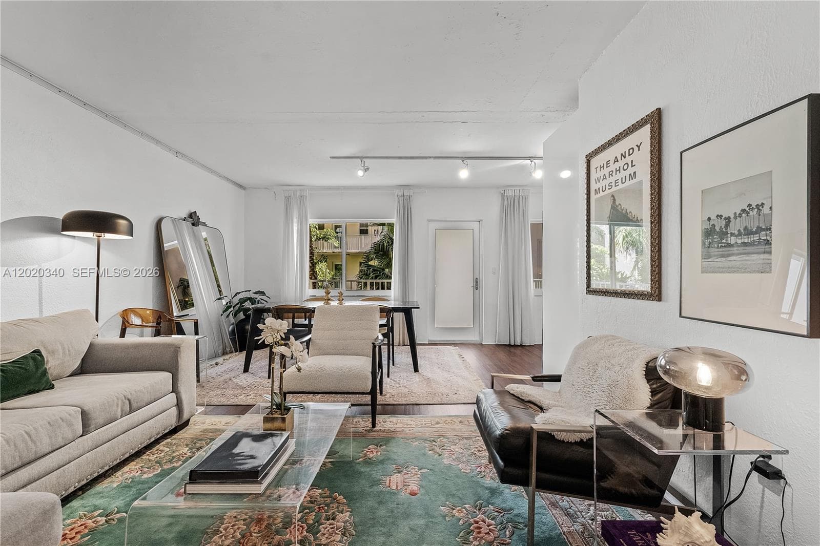 A bright, eclectic living space with white walls and track lighting, featuring a mix of vintage and modern furnishings — a light grey sofa, a dark leather chair, a transparent acrylic coffee table, and layered floral rugs. An Andy Warhol Museum poster and a palm tree photograph add artistic character to the walls, while a large leaning mirror and floor lamp bring depth to the room. A dining area with a dark table is visible toward the back, with natural light streaming through sheer curtains.
