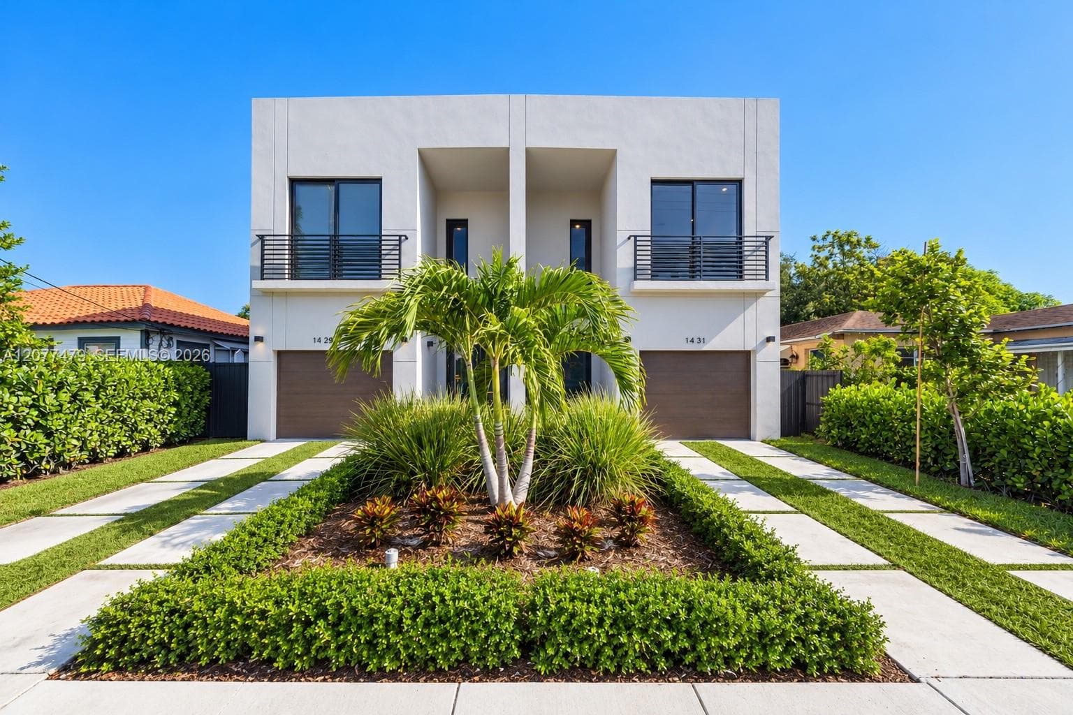 Modern two-story duplex exterior featuring a smooth stucco facade, two separate attached garages, minimalist metal balcony railings, and a concrete paver driveway