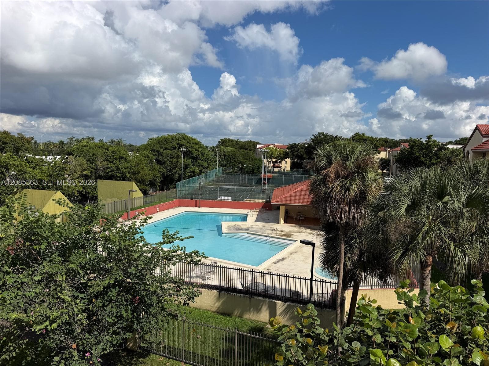 Outdoor recreational area featuring an in-ground swimming pool, covered pavilion, fenced tennis courts, and mature palm trees