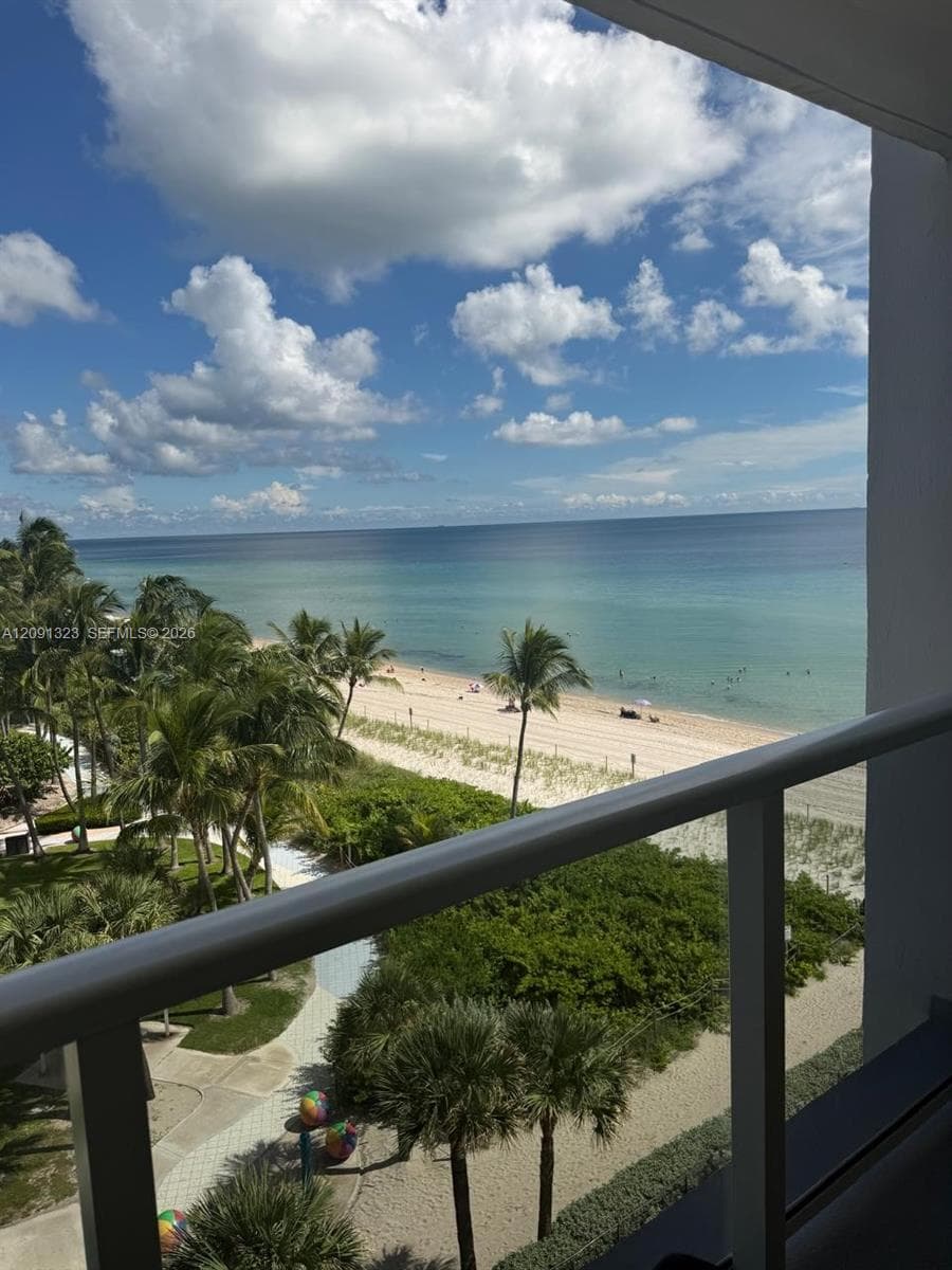 Private balcony featuring a metal railing, overlooking a sandy beach and oceanfront landscape with mature palm trees