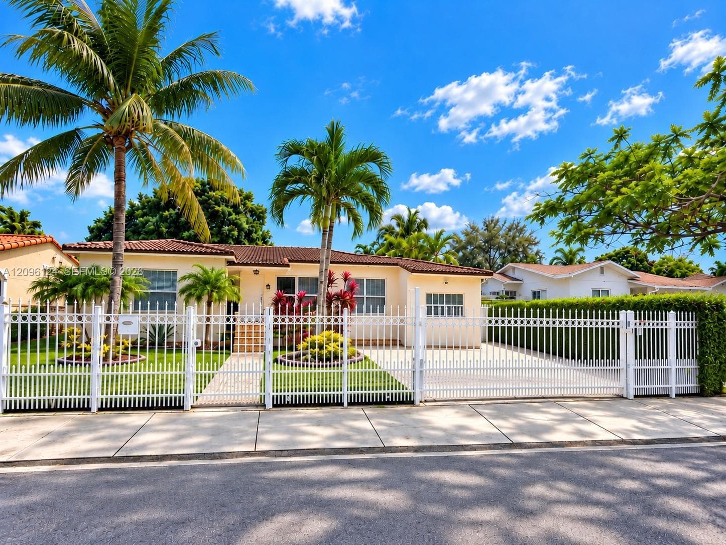 Single-story residence featuring a tile roof, light-toned exterior, gated perimeter fencing, paved driveway, and mature palm landscaping