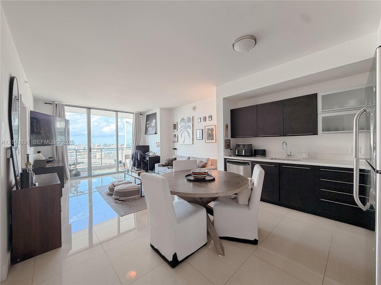 Open-concept interior featuring large-format tile flooring, kitchen with dark cabinetry, upper glass-front cabinets, and floor-to-ceiling glass sliding doors