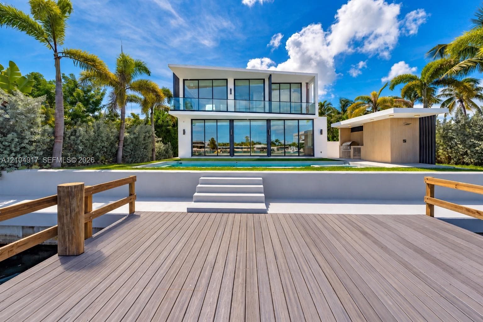 Waterfront property featuring a wood-finish dock with timber railings, an integrated in-ground swimming pool, a minimalist two-story residence with floor-to-ceiling glass windows, and a detached exterior kitchen structure