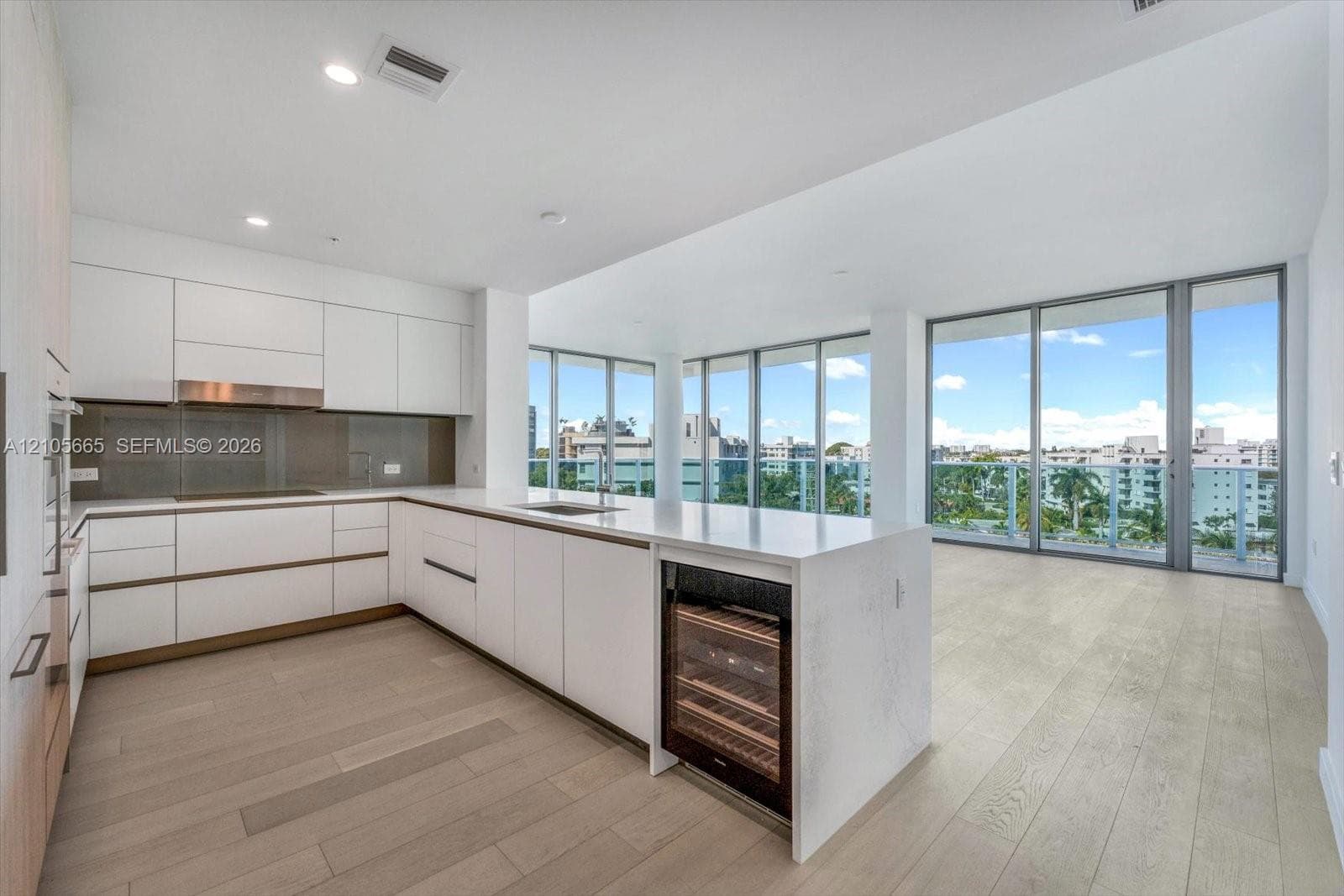 Contemporary kitchen featuring white flat-panel cabinetry, a stone-surfaced island with an integrated wine cooler, wood-finish flooring, and floor-to-ceiling glass panel walls