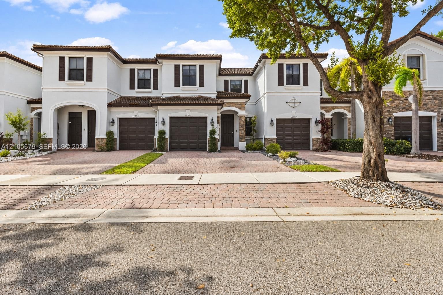 Multi-story exterior featuring light-toned stucco walls, decorative stone veneer accents, dark-finish carriage-style garage doors, tile roofing, and paver driveways