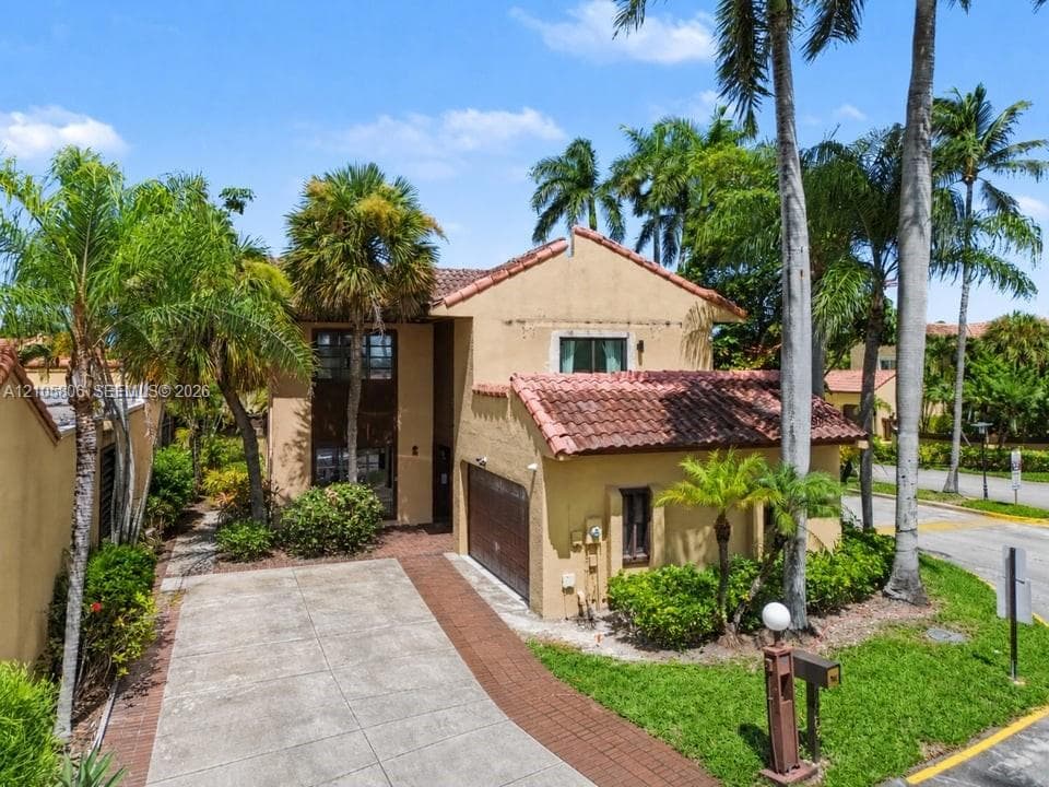 Two-story stucco exterior featuring a multi-pitched clay tile roof, attached garage, concrete driveway with brick paver border, and mature landscaping