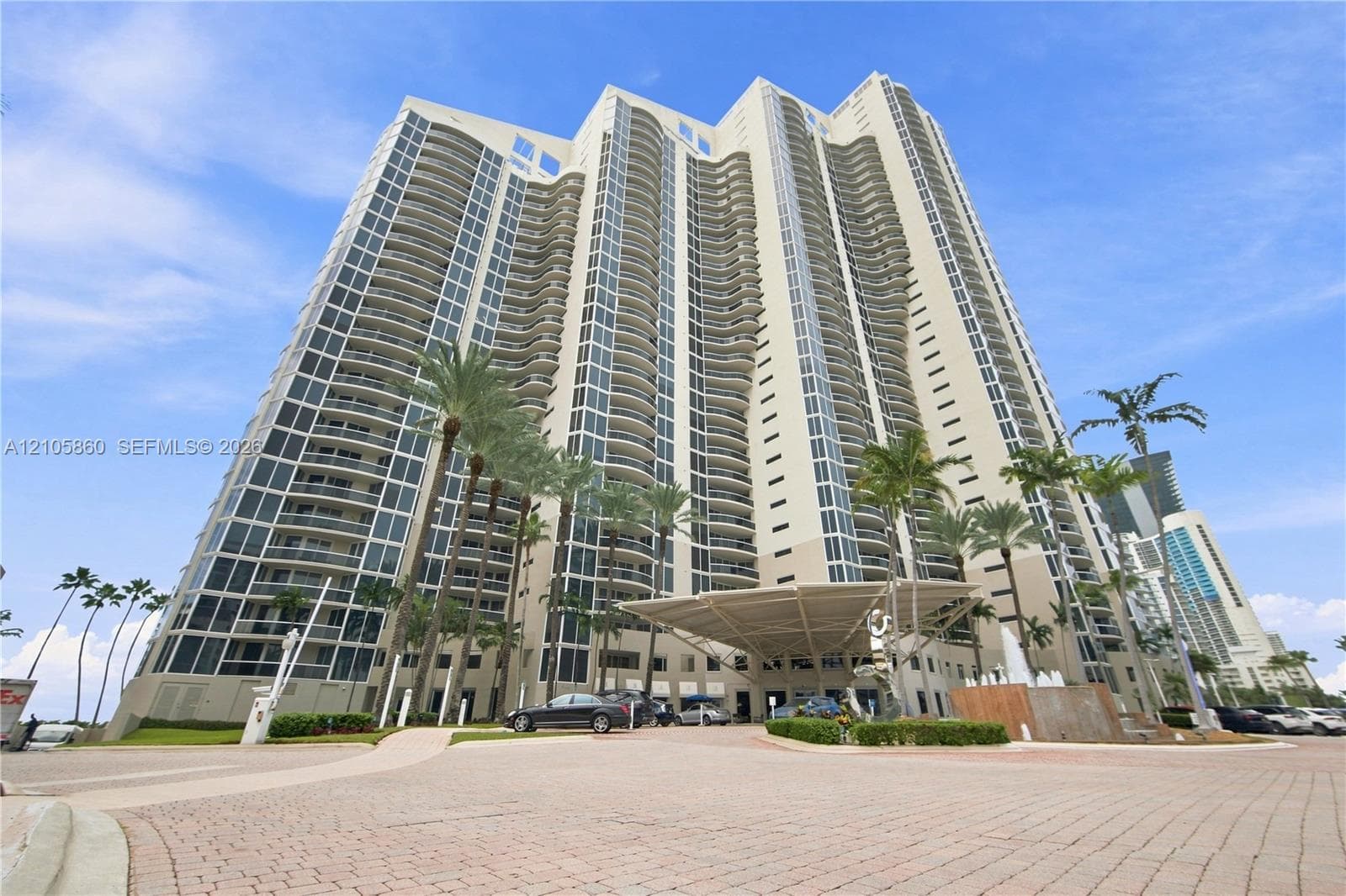 High-rise residential tower featuring a concrete facade, floor-to-ceiling windows, private balconies, a porte-cochère entry, and landscaped grounds with palm trees