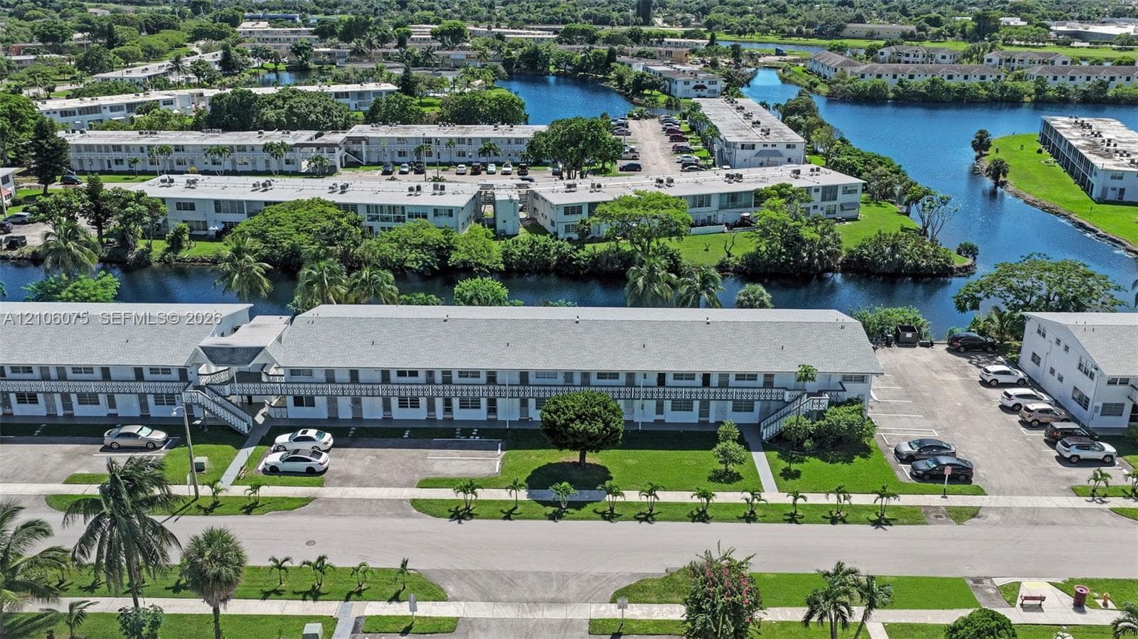 Multi-story building featuring exterior staircases, designated surface parking, mature palm trees, and waterfront grounds