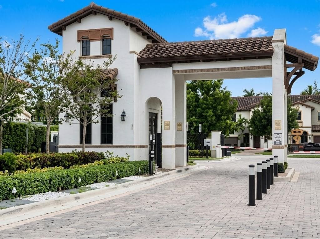 Gated entrance featuring a stucco guardhouse, clay tile roof, paver driveway, and controlled-access barrier arm