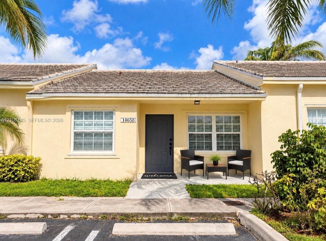 Single-story exterior featuring a light-toned stucco facade, dark-finish entry door, low-slope roof, and concrete patio