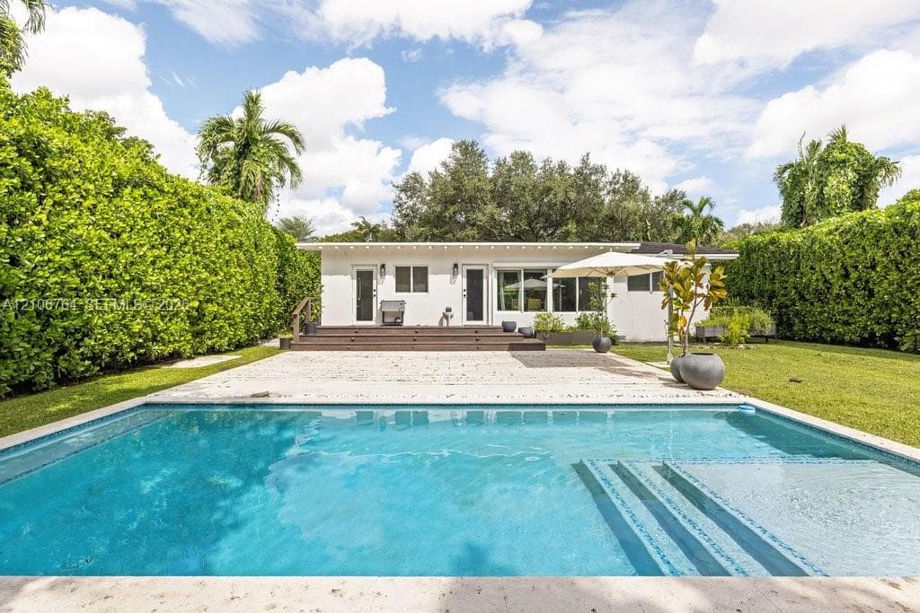Outdoor area featuring a swimming pool with integrated steps, stone-finish pool deck, elevated wood-finish deck, and surrounding manicured privacy hedges