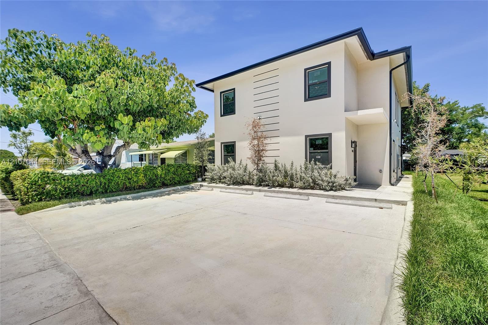 Two-story exterior featuring light-toned stucco siding, dark-trimmed windows, a concrete parking area, and mature landscaping