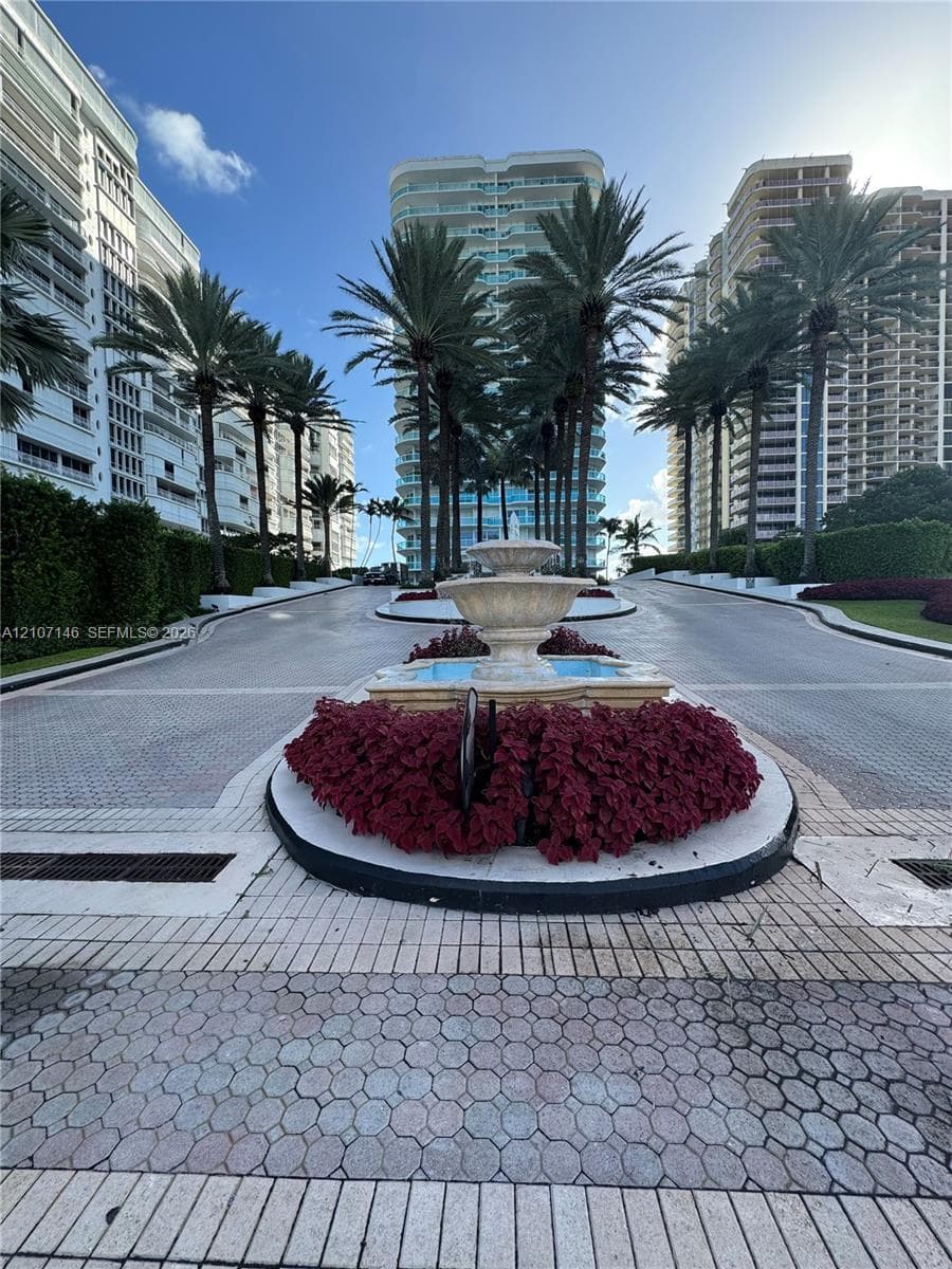 High-rise residential building with balcony glass railings, paved motor court with central fountain, private tennis court, and mature tropical landscaping