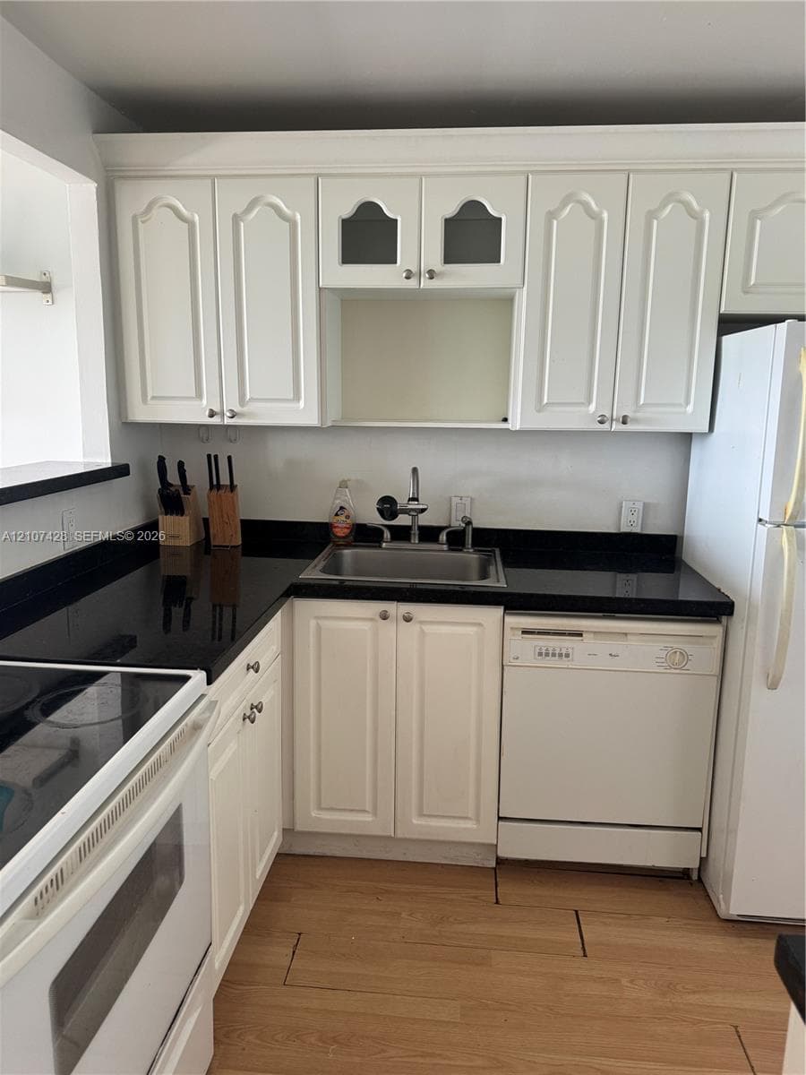 Kitchen featuring white cabinetry with glass-panel accents, dark countertops, a stainless steel undermount sink, and wood-finish flooring