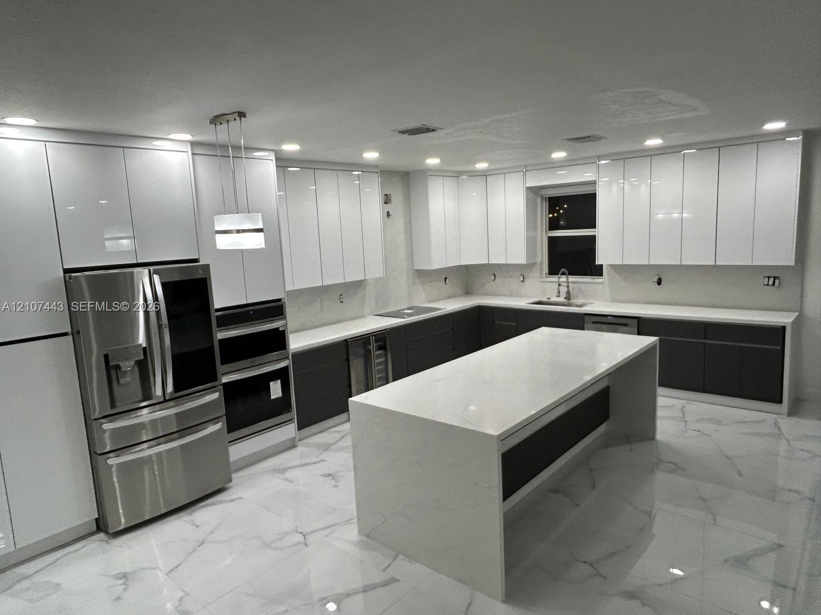 Kitchen featuring high-gloss white upper cabinetry, dark lower cabinetry, a large central island with a stone-finish waterfall countertop, stainless steel appliances, and large-format tile flooring