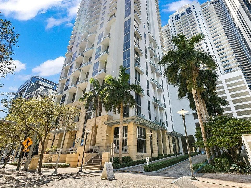 High-rise residential exterior featuring a white facade, staggered private balconies, multi-story glass windows, stone-paved driveway, and mature palm tree landscaping