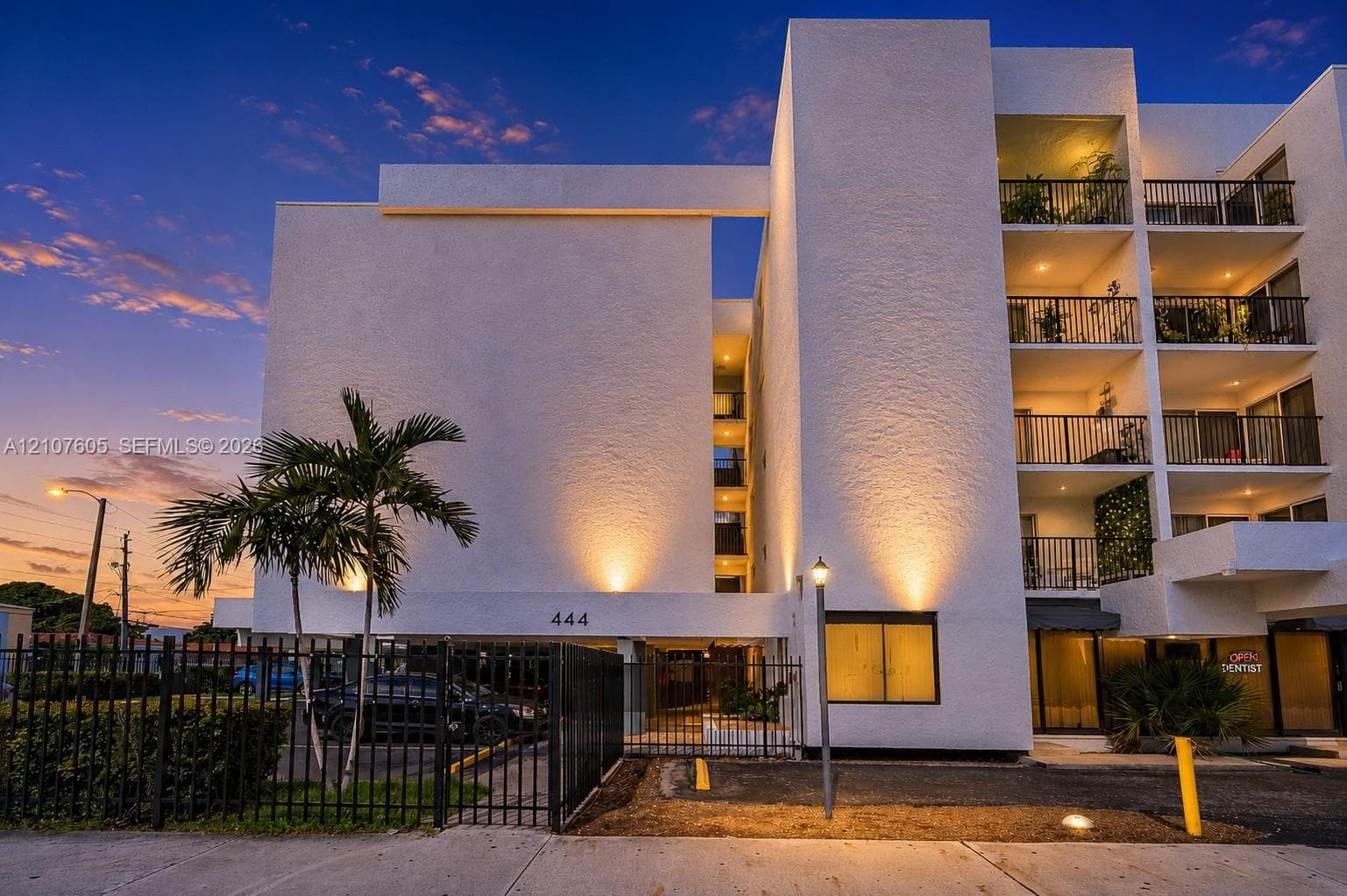 Multi-story stucco exterior featuring stacked private balconies, black metal railings, and a gated parking entrance
