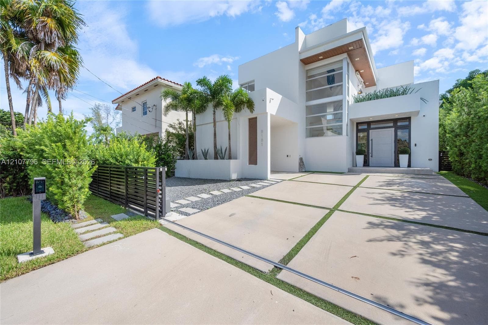 Modern architectural exterior featuring a multi-level white facade, geometric concrete paver driveway, full-height glass window wall, and integrated wood-finish ceiling accents
