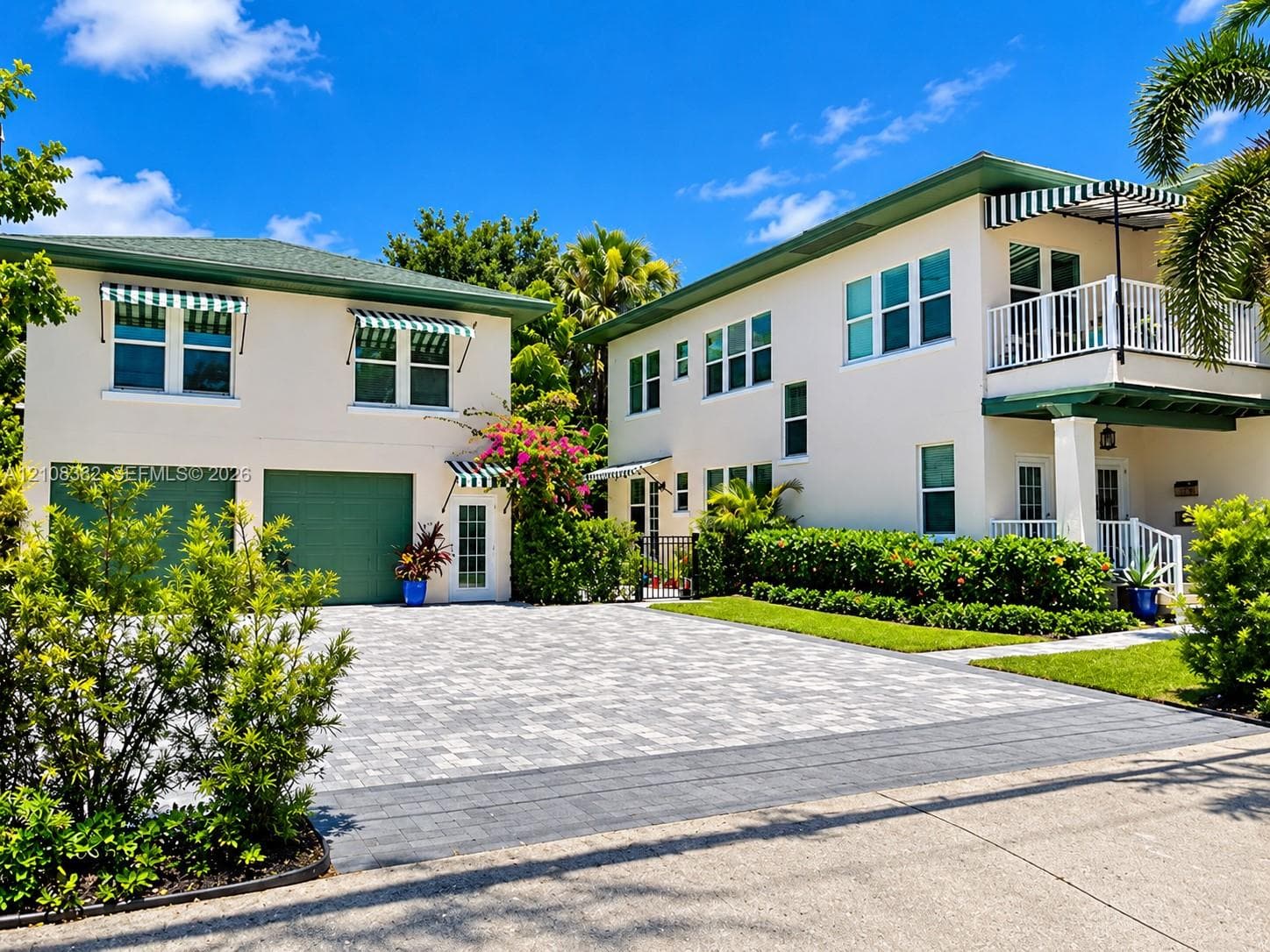 Two-story exterior featuring multi-tone paver driveway, green-tinted garage doors, striped fabric awnings, a second-story balcony with white railing, and structured landscaping