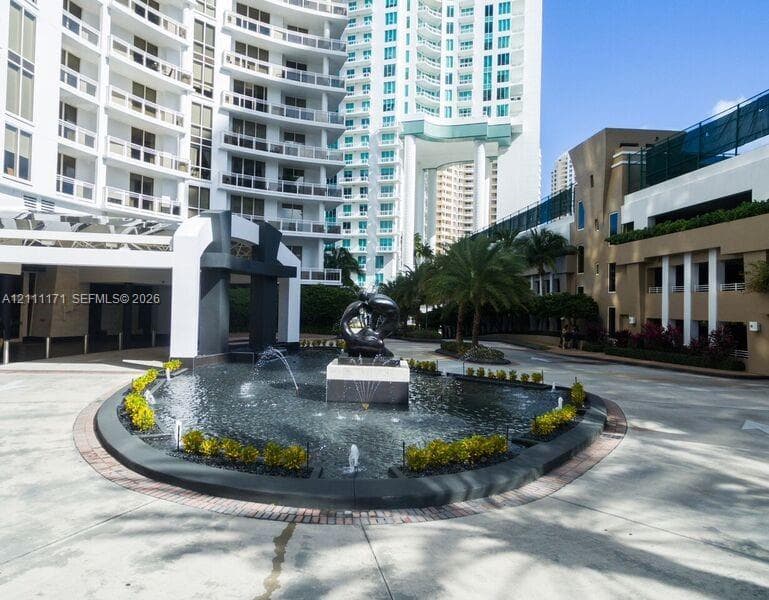 Circular motor court featuring a tiered stone fountain, central bronze-toned sculpture, concrete paver accents, and structured landscaping